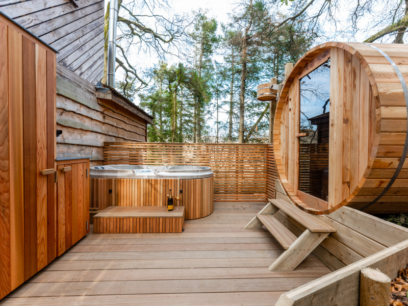 Outdoor spa area with a wooden deck featuring a round wooden sauna, a modern hot tub, and wooden cabinets. Surrounded by a wooden fence and tall trees in the background, this unique accommodation offers a natural and serene atmosphere perfect for your stay at Ravendere Treehouse.