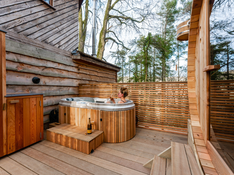 A person relaxes in a wooden hot tub on the deck of the Ravendere Treehouse, surrounded by a wooden privacy fence. Trees visible in the background enhance the natural ambiance. A bottle of beer and a glass sit nearby, adding to the rustic charm of this cozy cabin retreat.