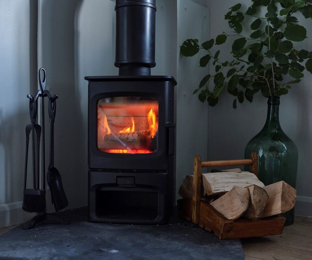 A cozy wood-burning stove with a fire inside sits on a black hearth in the Devon cottage. Beside it, a basket of firewood rests on the wooden floor. A green vase with leafy branches is tucked in the corner, adding a touch of nature to this Sleepy Owl retreat.