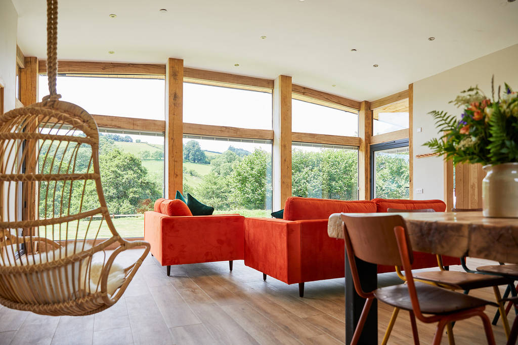 A spacious living room with large windows offering a view of Burrows Dolassey's lush green hills. It features orange sofas, a wooden swing chair, a dining table adorned with flowers, and an inviting atmosphere that radiates warmth.