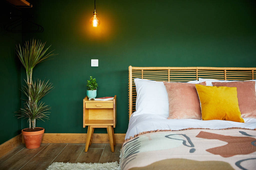 A cozy Burrows bedroom with a green accent wall, a wooden headboard, and a neatly made bed featuring pillows in white, pink, and yellow. A Dolassey nightstand with a plant and book sits beneath a glowing pendant light. A potted plant is in the corner.