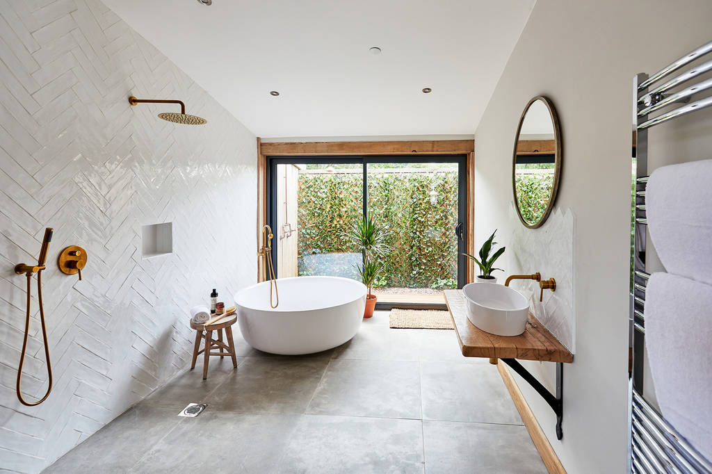 A modern bathroom featuring a freestanding bathtub, walk-in shower with gold fixtures, and a wooden vanity with a round mirror. Large glass doors offer a view of the Burrows Dolassey garden. The room has white herringbone tiles and gray floor tiles.