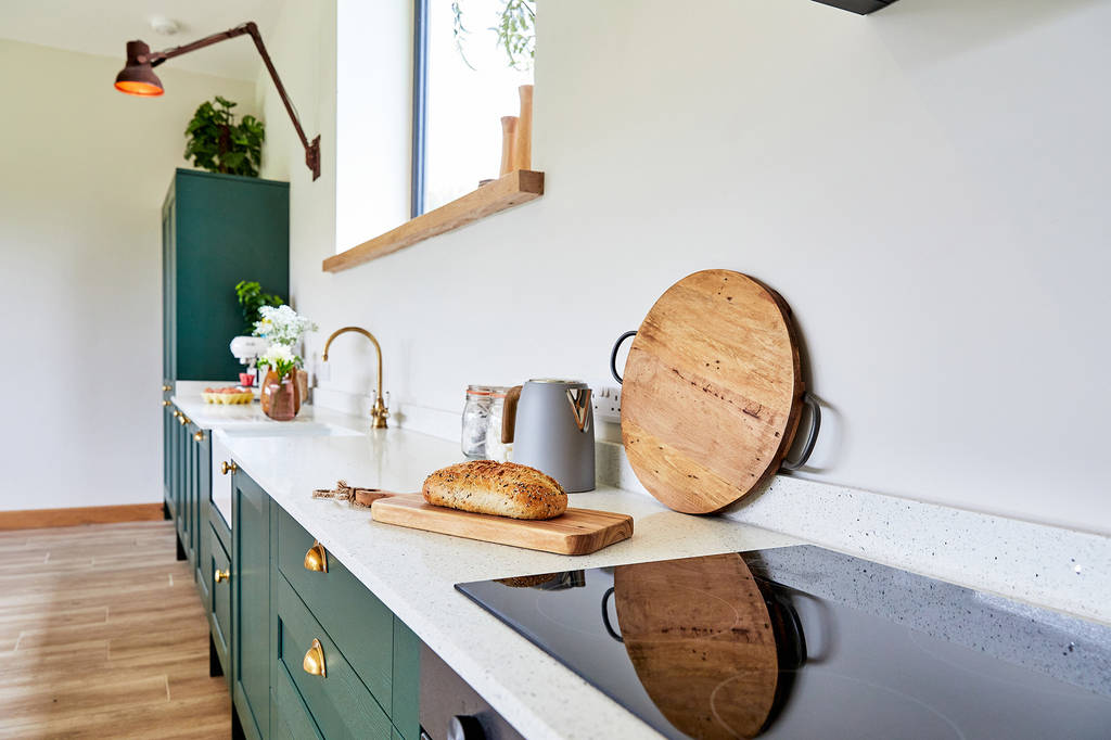 A modern kitchen with Burrows green cabinets and wooden accents. The countertop features a loaf of bread on a cutting board, a Dolassey wooden serving board, a toaster, and decorative plants. The room is well-lit with natural light from a window.