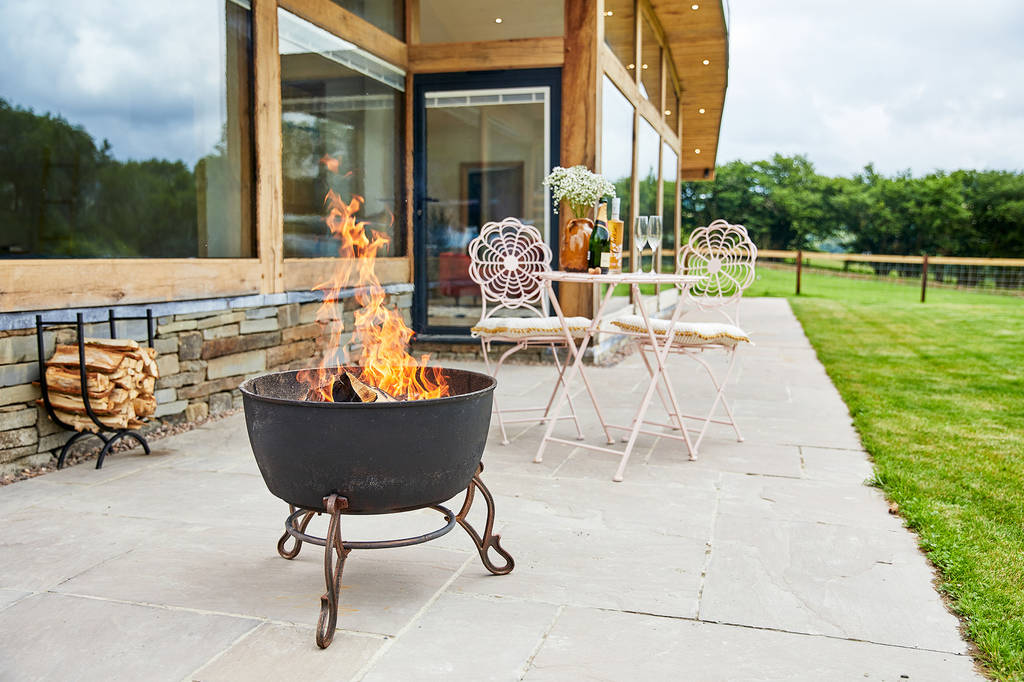 A cozy outdoor patio features a metal fire pit with flames, a rustic table set with two ornate chairs, and a view of a grassy lawn. A stack of firewood is nearby, while the patio is bordered by Dolassey's modern glass-walled building.