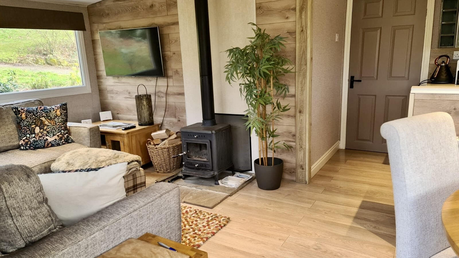 A cozy living room reminiscent of woodland escapes, featuring a wood-burning stove, a TV, and a comfy couch. A small plant adds charm, while a notepad rests invitingly on the wooden coffee table—perfect for jotting down thoughts in this serene lodge-like setting.