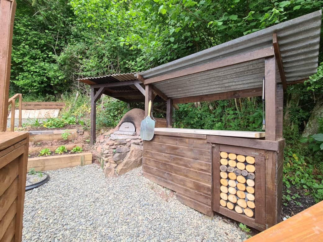 An outdoor pizza oven with a corrugated metal roof and wood storage section, nestled in a garden surrounded by lush greenery—a perfect addition for those seeking woodland escapes.