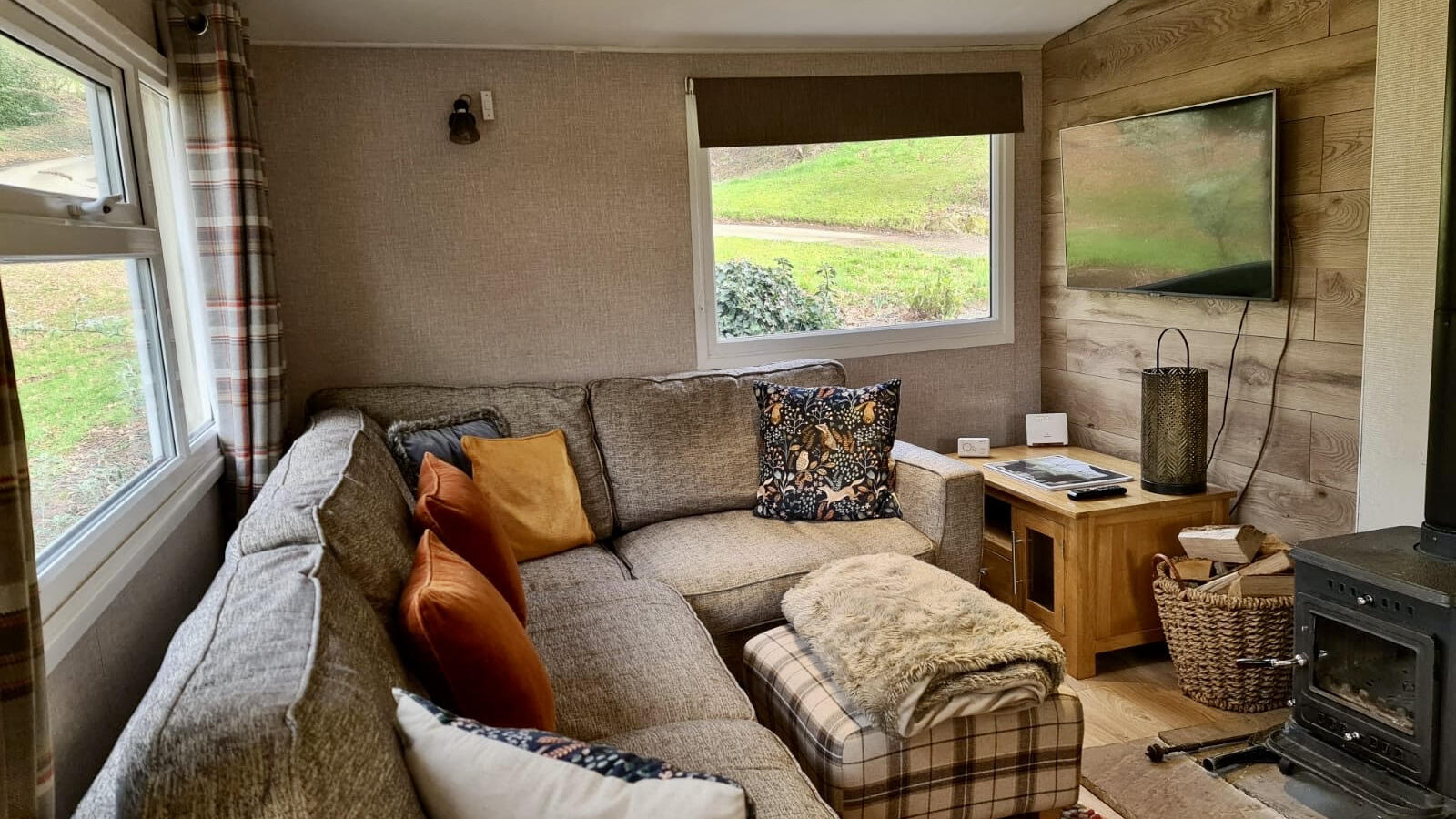 Cozy living room with a corner sofa, plaid ottoman, cushions, wall-mounted TV, and wood-burning stove. Large windows overlook lush woodland greenery, creating the perfect escape from everyday life.