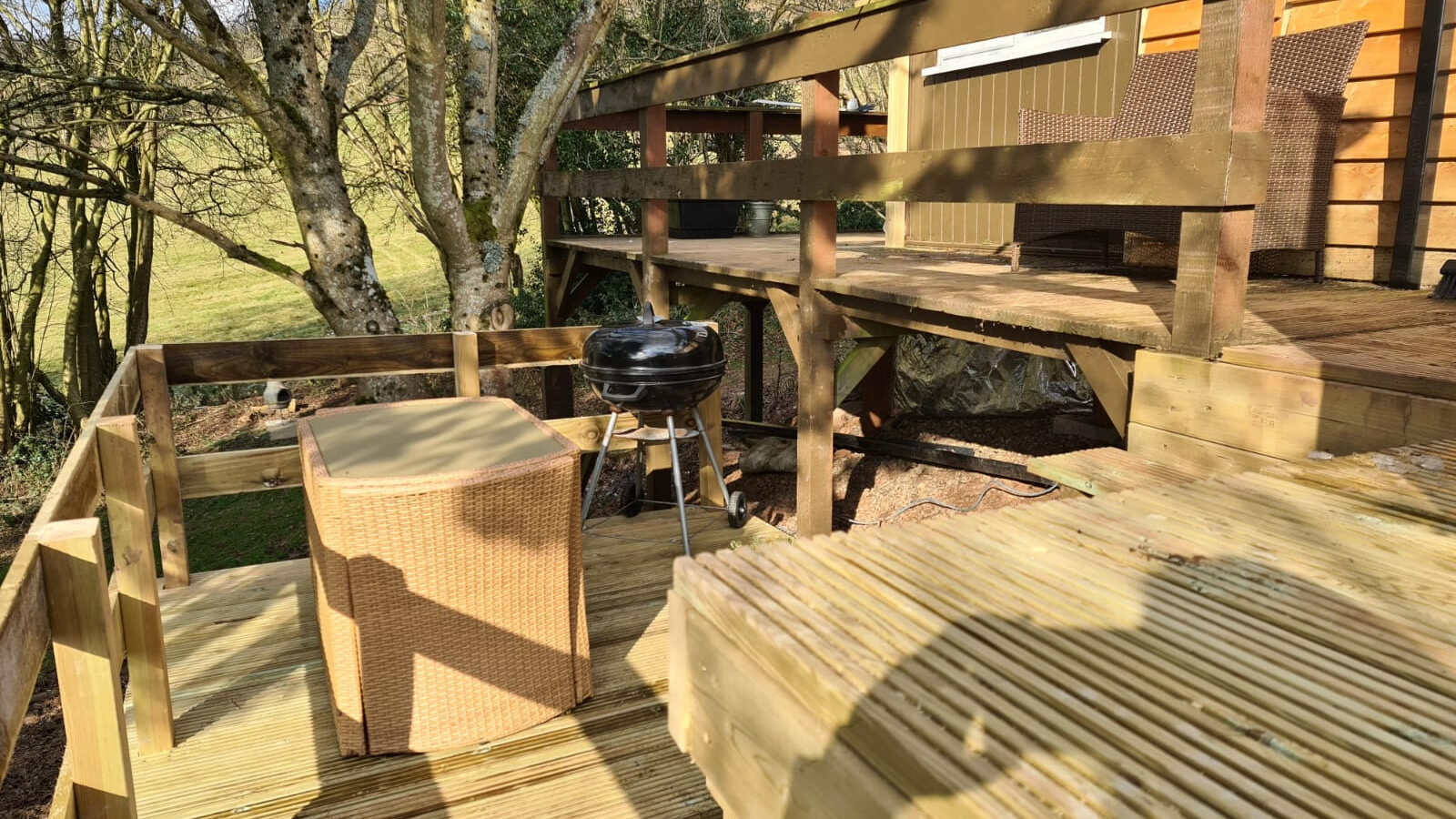 Nestled amidst the woodland, this wooden deck offers the perfect escape with its wicker furniture, a small charcoal grill, and surrounding trees under a bright sky.