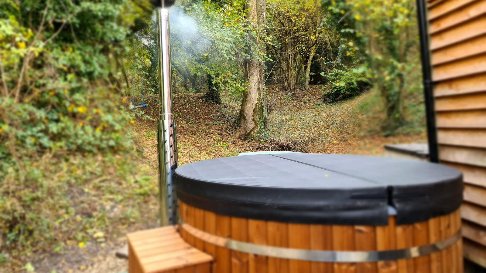 A wooden hot tub with a covered lid and chimney emitting smoke sits serenely in the wooded area next to a rustic treehouse, perfect for star gazing on clear nights.