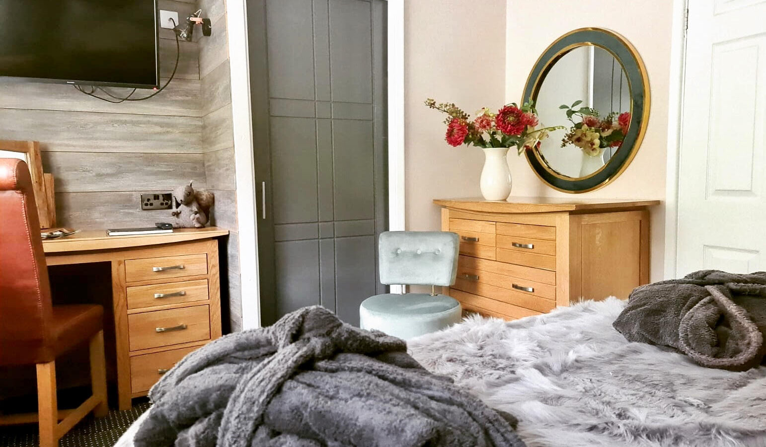 Escape to a serene woodland lodge featuring a cozy bedroom with a bed draped in a grey blanket and robes in the foreground. A chair, desk, dresser, TV, and mirror adorned with flowers complete the tranquil setting.