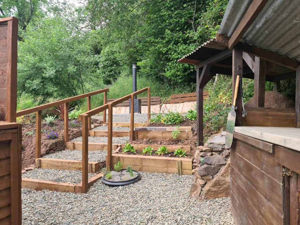 A terraced garden with wooden steps, planters, and gravel paths offers a peaceful woodland escape. A metal trowel hangs on a nearby wooden structure, echoing the charm of rustic lodges nestled in nature's embrace.