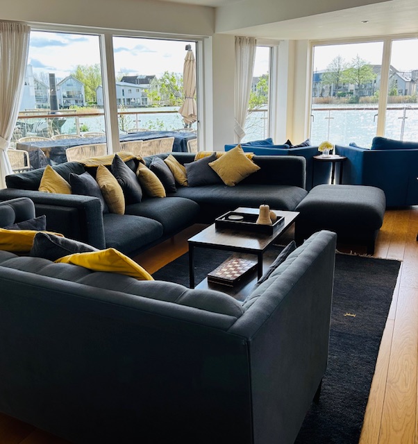 A spacious living room on Lamplight Island features dark blue sofas, yellow and blue pillows, and large windows overlooking a serene lake.