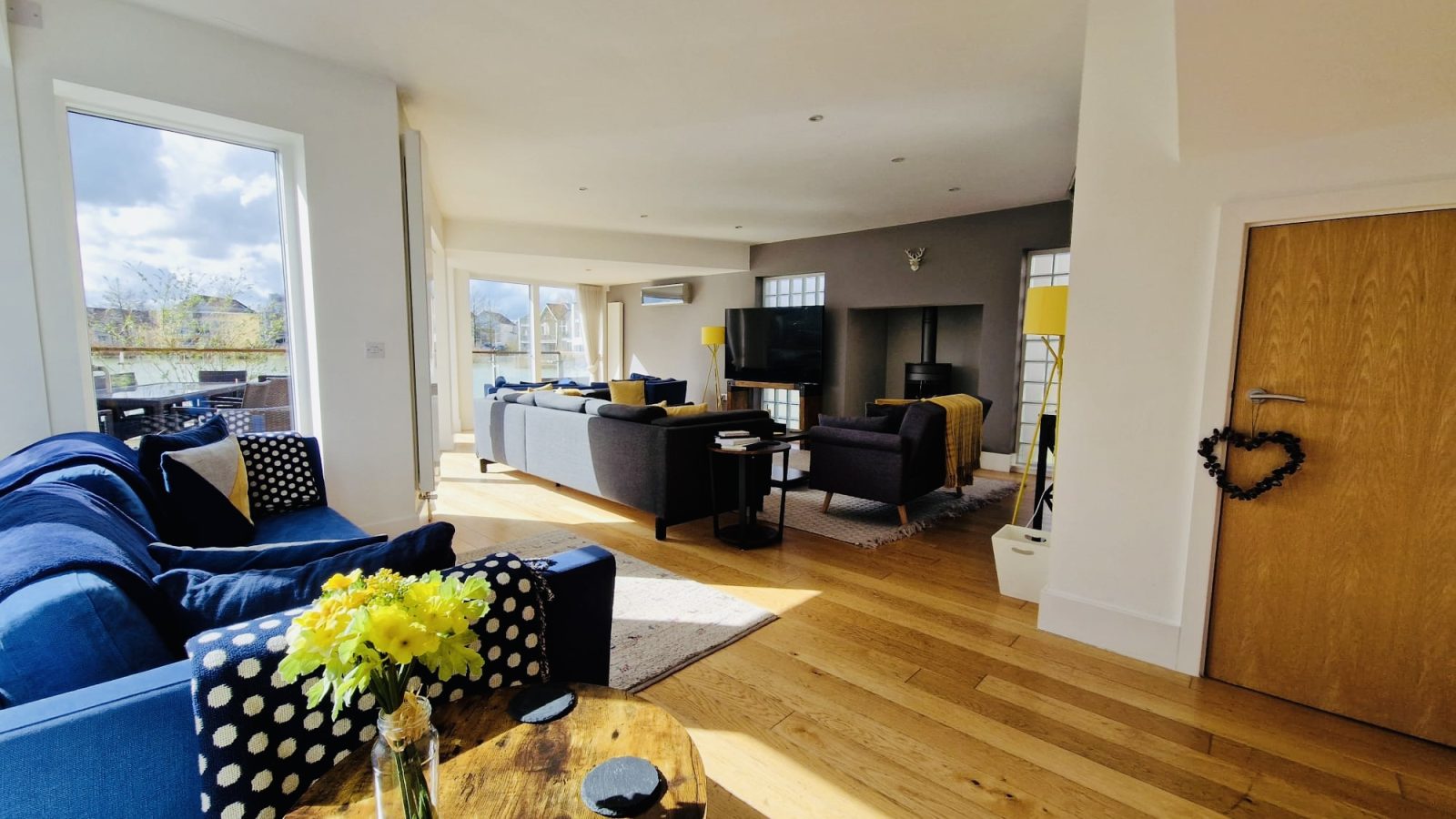 The bright living room features wooden flooring, blue sofas, and yellow accents, illuminated by the soft glow of a Lamplight Island fixture. Daffodils grace the table, perfectly complementing the serene view of the outdoors.