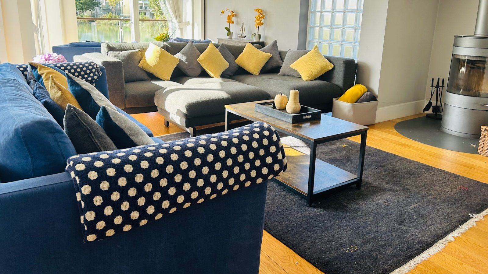 A living room featuring a blue and gray sectional sofa with yellow cushions, a black rug, and a coffee table is beautifully illuminated by the soft glow of lamplight. The fireplace adds warmth, creating an inviting island of comfort.