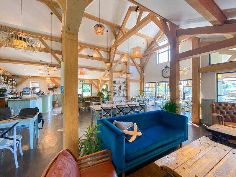 Lamplight Island-themed cafe interior with wooden beams, blue sofa, and tables. Natural light floods through large windows.