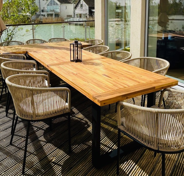 The outdoor dining area on Lamplight Island features a long wooden table surrounded by wicker chairs, perfectly set on a deck with views of the tranquil water and charming houses.