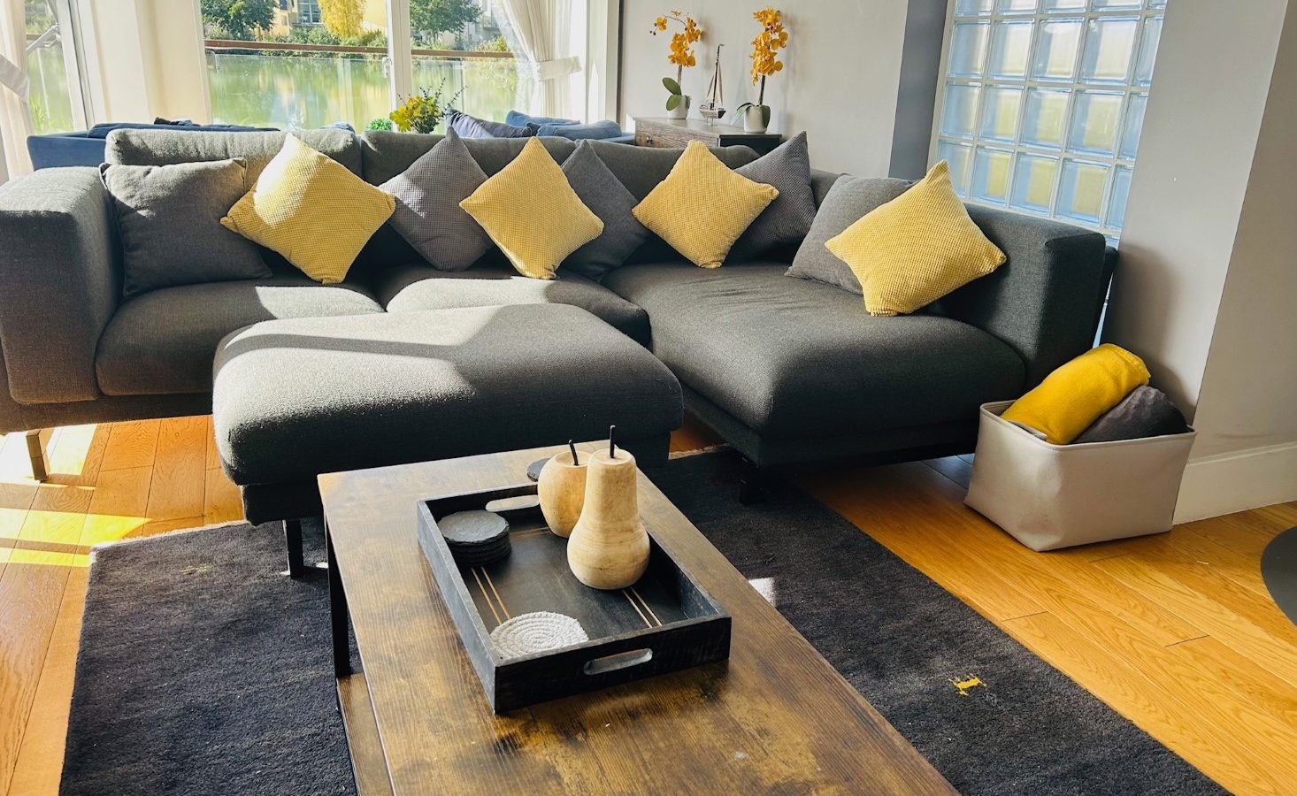 A modern living room features a gray sectional sofa adorned with yellow pillows, a wooden coffee table at its center, and the gentle glow of lamplight casting a warm ambiance alongside the sunlight streaming through the window.