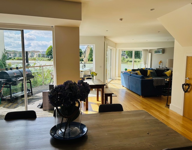 Bright open-plan living space with a dining table, blue sofa, and large windows overlooking Lamplight Island's patio, complete with grills and lush greenery.