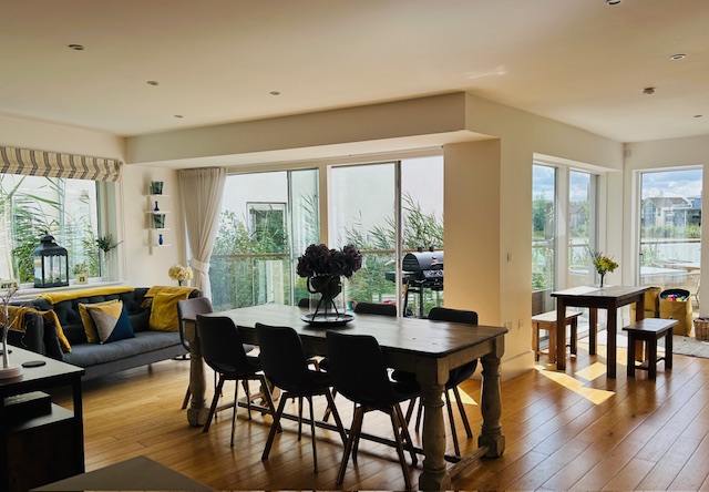 Bright dining room featuring a wooden table, black chairs, and a cozy sofa with yellow cushions. Large windows illuminate the space and highlight the charm of Lamplight Island, complemented by a quaint bench table.