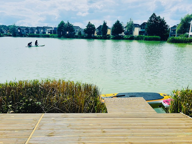 Two people paddleboarding on a serene lake, with the charming houses and lush trees of Lamplight Island in the background and a rustic wooden dock in the foreground.