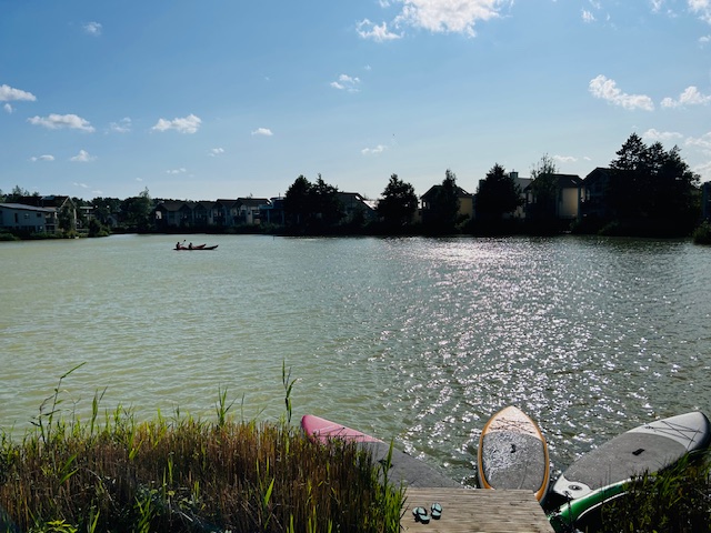 Two people paddle a kayak on a sunlit lake near Lamplight Island, with several paddleboards in the foreground and houses in the background.