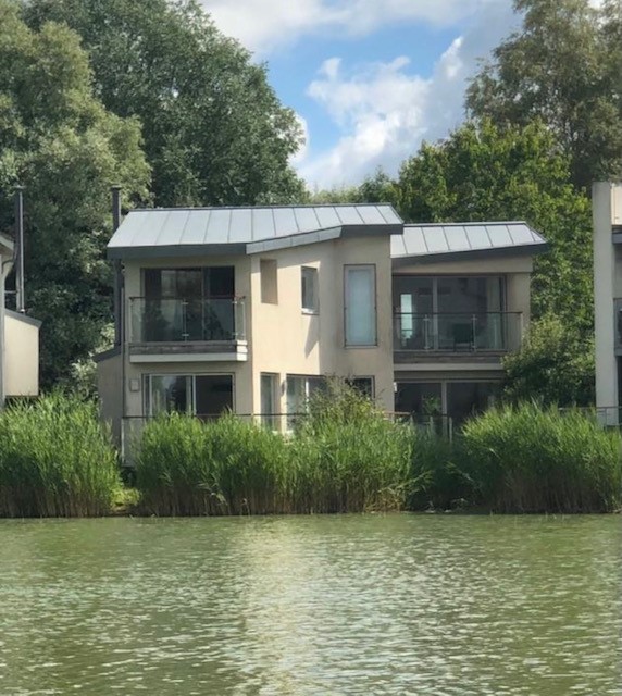 Nestled on Lamplight Island, this modern two-story house features large windows and a sleek metal roof. It's beautifully situated near a tranquil body of water, enveloped by trees and reeds.