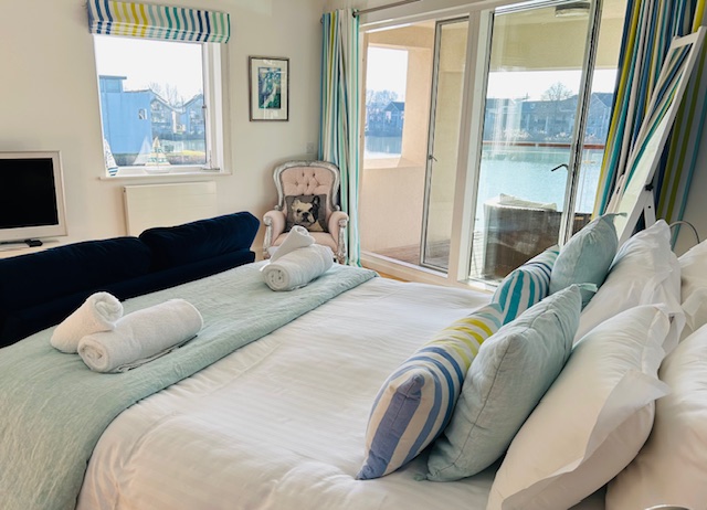 Bright bedroom with a large bed, striped pillows, and towels. Glass doors open to a balcony overlooking the serene waters of Lamplight Island.