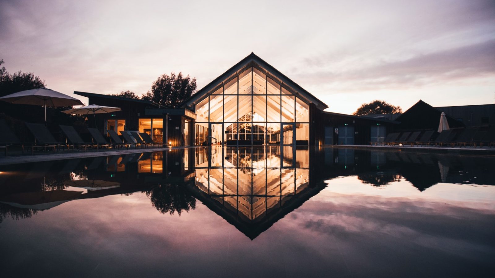 A modern building with a glass facade is illuminated from within, reflected perfectly on the calm water of a pool in the foreground. It's dusk on Lamplight Island, where the sky is a soft gradient of pink and purple, enhancing the tranquil atmosphere.