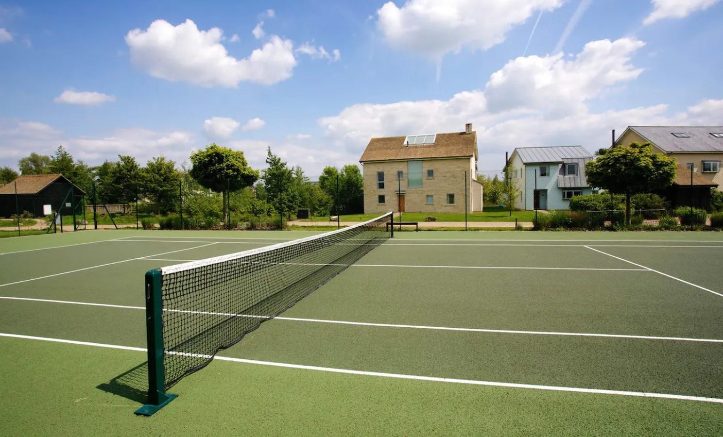 A tennis court with a net in the foreground is nestled on Lamplight Island, surrounded by lush green trees and bushes. In the background, modern two-story houses stretch toward a blue sky dotted with scattered clouds.