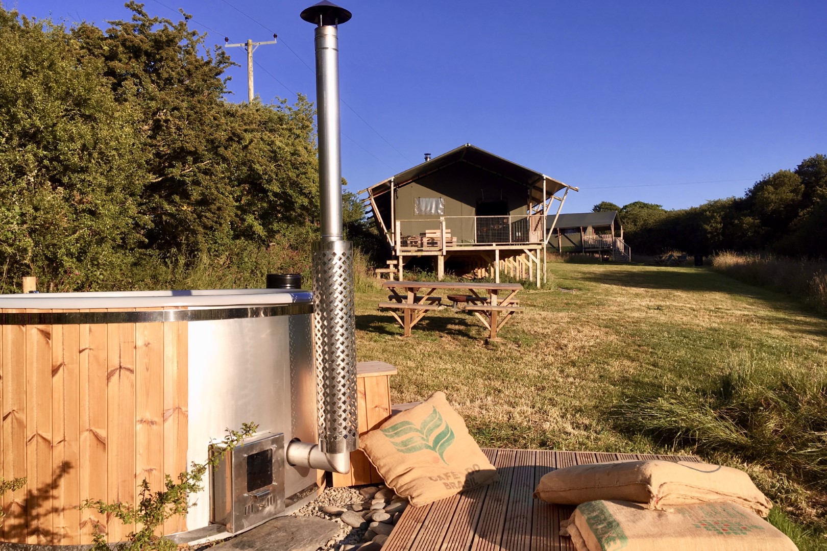 Wooden hot tub with chimney on grassy area, picnic tables, and tent structure—ideal for cheap breaks with hot tubs in the UK.