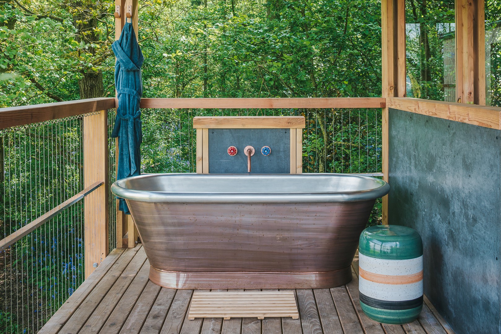 Discover the charm of UK stays with outdoor baths, featuring a copper-finished bathtub nestled amidst lush greenery on a wooden deck, complete with a cozy towel and an inviting decorative stool.