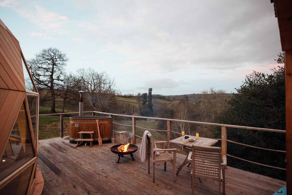 Rural deck scene at The Nova Dome with wooden chairs, fire pit, hot tub, and a countryside view under a cloudy sky.