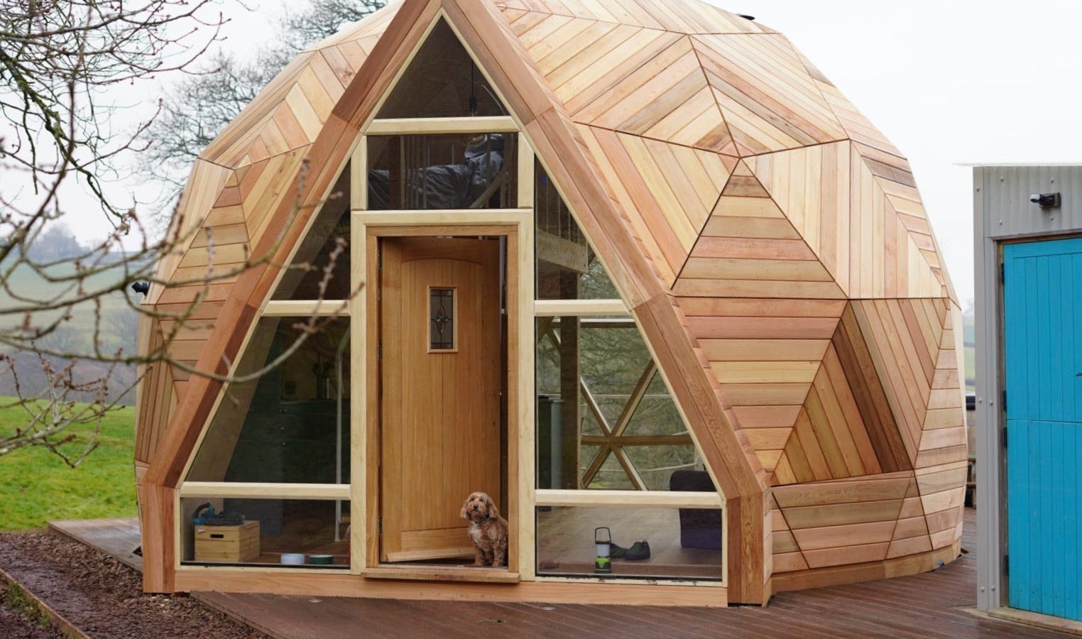 The Nova Dome at The Fold: A geodesic wooden cabin with a central door on a deck, where a small dog sits at the entrance.