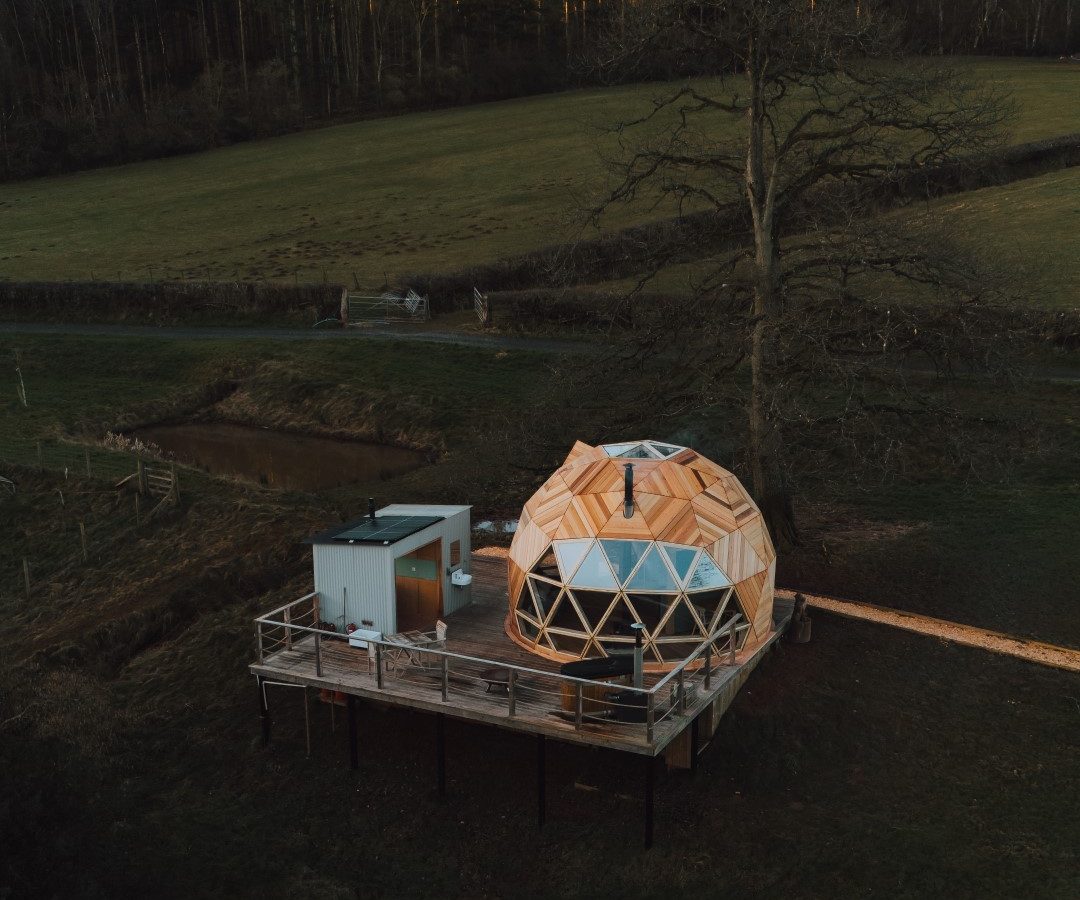 The Nova Dome at The Fold features a geodesic cabin with a wooden deck nestled in lush greenery under a clear sky.