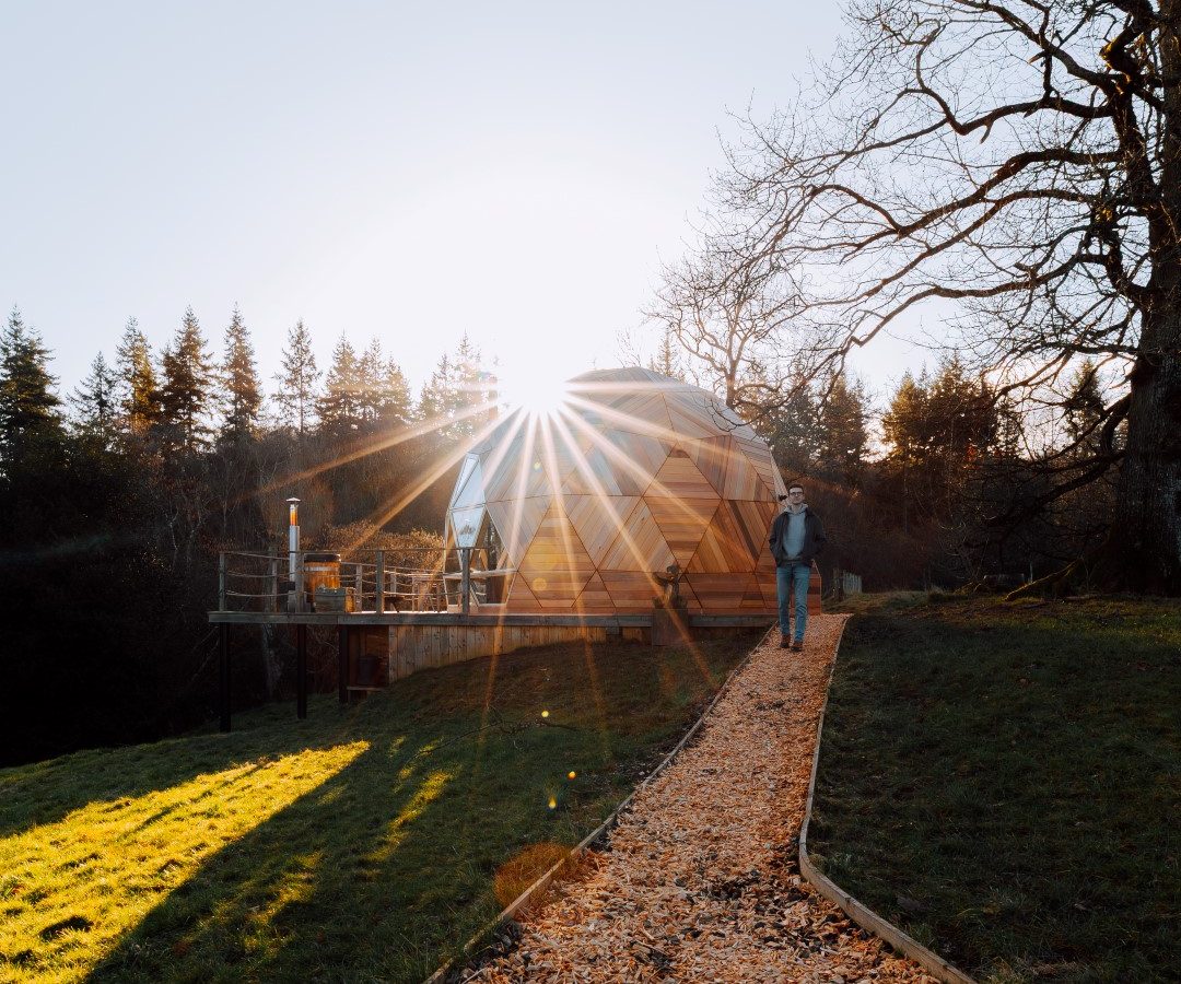 Walking on a wood chip path, they approach The Nova Dome at The Fold, surrounded by trees and a sunburst glow.