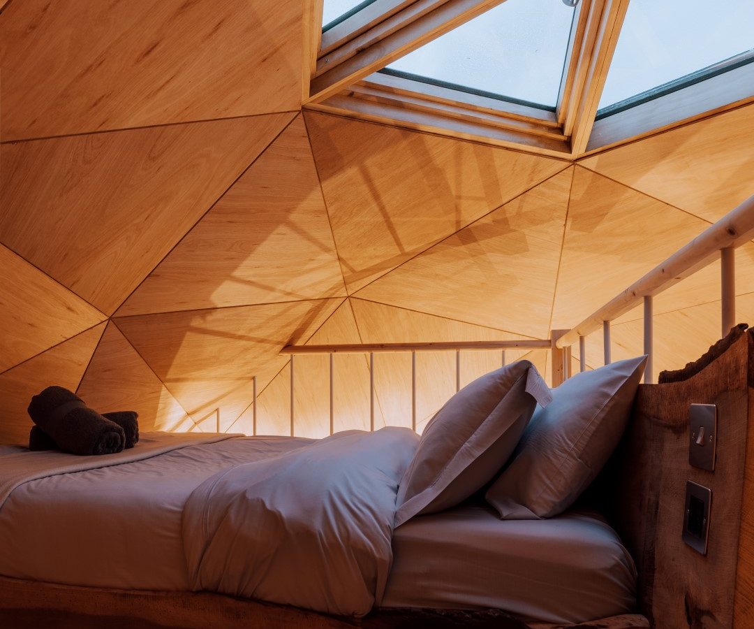 The Nova Dome at The Fold: A wood-paneled bedroom with a skylight, sunlight streaming over the neatly arranged bed and towels.