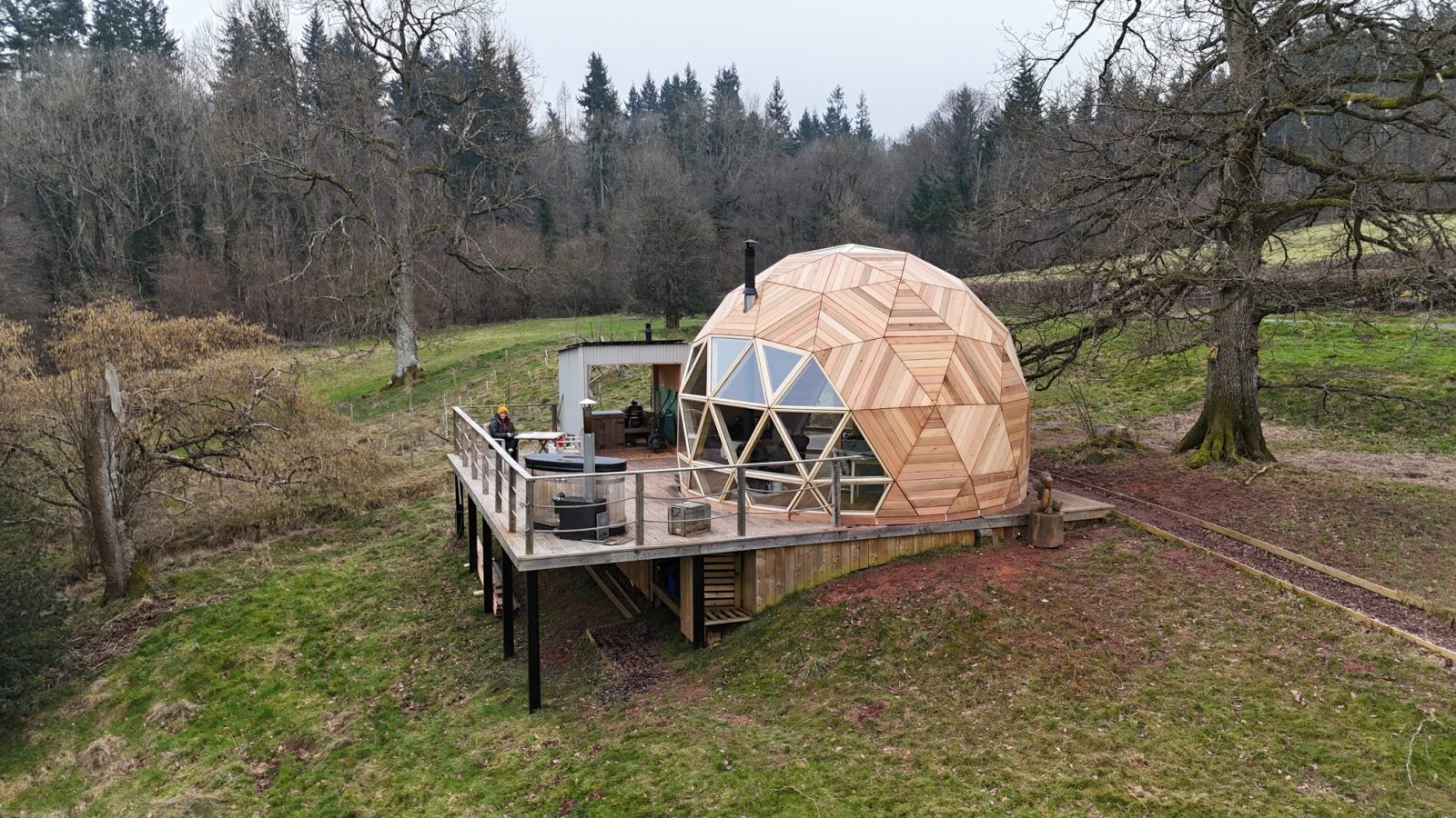 The Nova Dome at The Fold is a geodesic cabin with a wooden deck, nestled amidst grass and wooded tranquility.