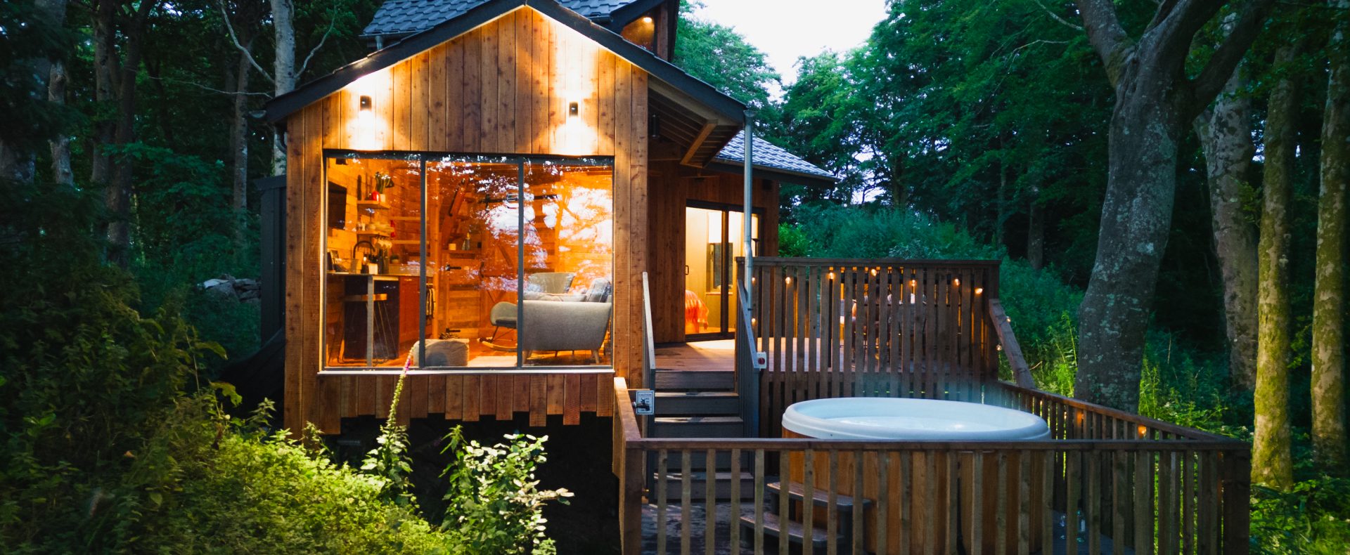 A treehouse with large glass windows, nestled among trees, offers a serene deck with a hot tub, perfect for adults-only breaks in the UK.