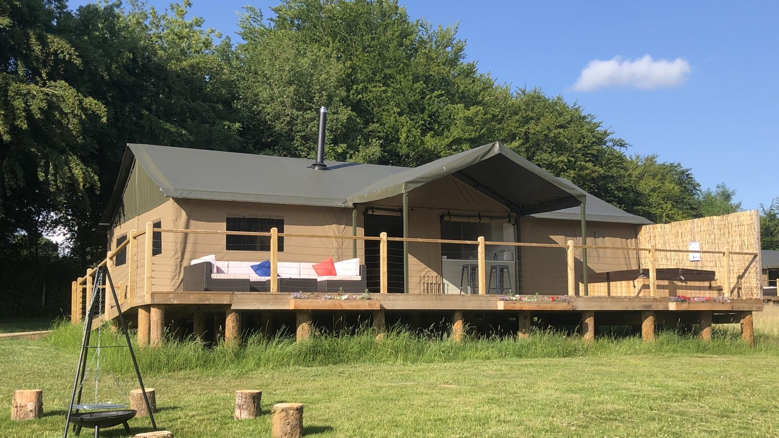 A glamping tent on a raised wooden platform with a shaded porch area, surrounded by the lush flora of nature. A line with colorful towels and rustic wooden seating create a cozy outdoor atmosphere under a clear blue sky.