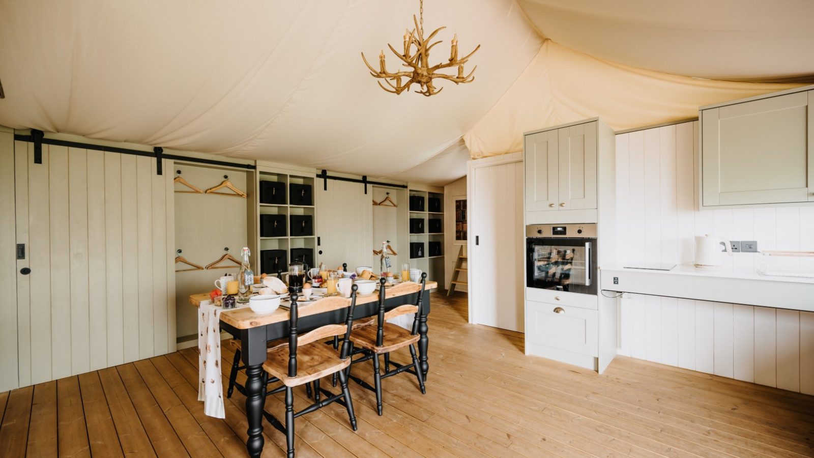 A rustic, cozy kitchen with wooden floors and cream walls. A wooden dining table is set with mugs, plates, and teapots, evoking a sense of flora and fauna. Shelving with hangers is in the background. A chandelier resembling antlers hangs from the ceiling.