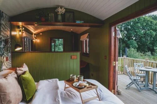 A cozy cabin bedroom with green walls features a bed adorned with plush pillows and a wooden tray holding breakfast. The room opens onto a wooden deck with a table and chairs, offering a view of lush greenery outside. Warm lighting enhances the serene ambiance, inviting you to log in to relaxation.