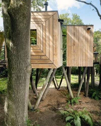 A modern treehouse made of wooden panels stands elevated on stilts among lush greenery. It features large square windows and angular design, blending contemporary architecture with natural surroundings. Sunlight filters through the trees, casting gentle shadows, offering users a serene retreat.