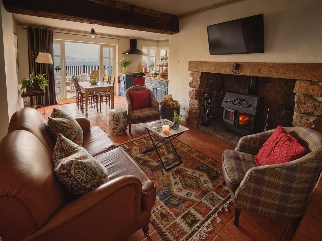 Cozy living room with a leather sofa, armchair, rug, and fireplace. The TV and dining area by the balcony offer a stunning view. Perfect for Devon stays with outdoor baths that add an extra touch of relaxation.