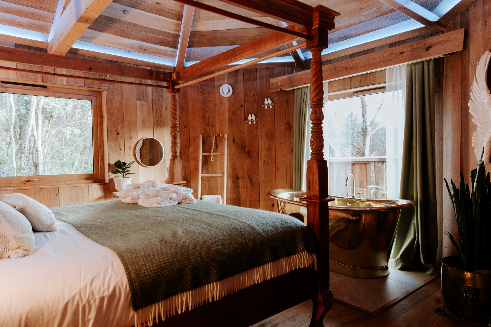 A cozy wooden bedroom with a large bed, gold freestanding bathtub by the window, and rustic decor offers a perfect retreat for those seeking unforgettable Devon stays.