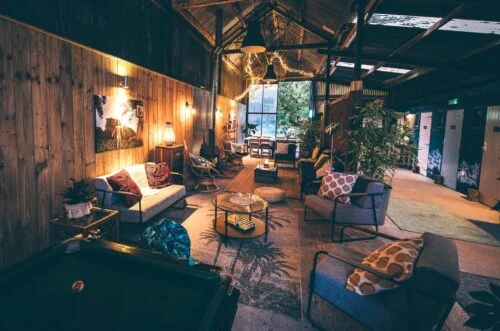 A cozy, rustic lounge area with wooden walls and soft lighting. The room features various seating options, including armchairs and couches, surrounding a pool table. Decorative throw pillows and plants add a touch of warmth. Log in to enjoy the comfort through an open doorway leading outside.