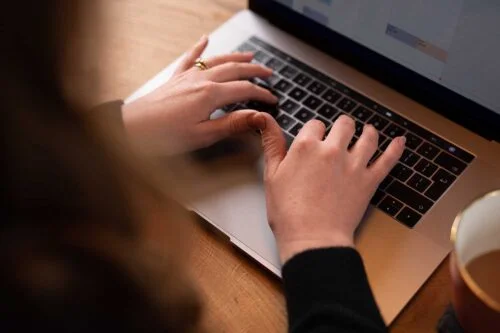 Close-up of a person's hands typing on a laptop keyboard, likely working on a login screen. The person is wearing a black long-sleeve top and has a ring on their right hand. A cup of coffee sits to the right of the laptop on a wooden table, with the screen partially visible.