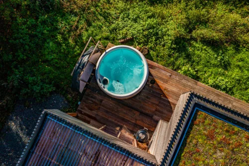 Aerial view of a wooden deck featuring a circular hot tub filled with bubbling water. Adjacent to the hot tub is a small table with a drink on it, inviting you to relax without needing to login. The deck is surrounded by vibrant green foliage, giving it a secluded and peaceful atmosphere.