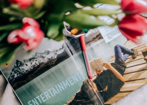 A magazine is open to an entertainment page featuring an image of a person lying on a wooden dock by a lake, reading a book. Snow-capped mountains are in the background. A red pen rests on the magazine, alongside user access notes, and red flowers are partially visible in the foreground.