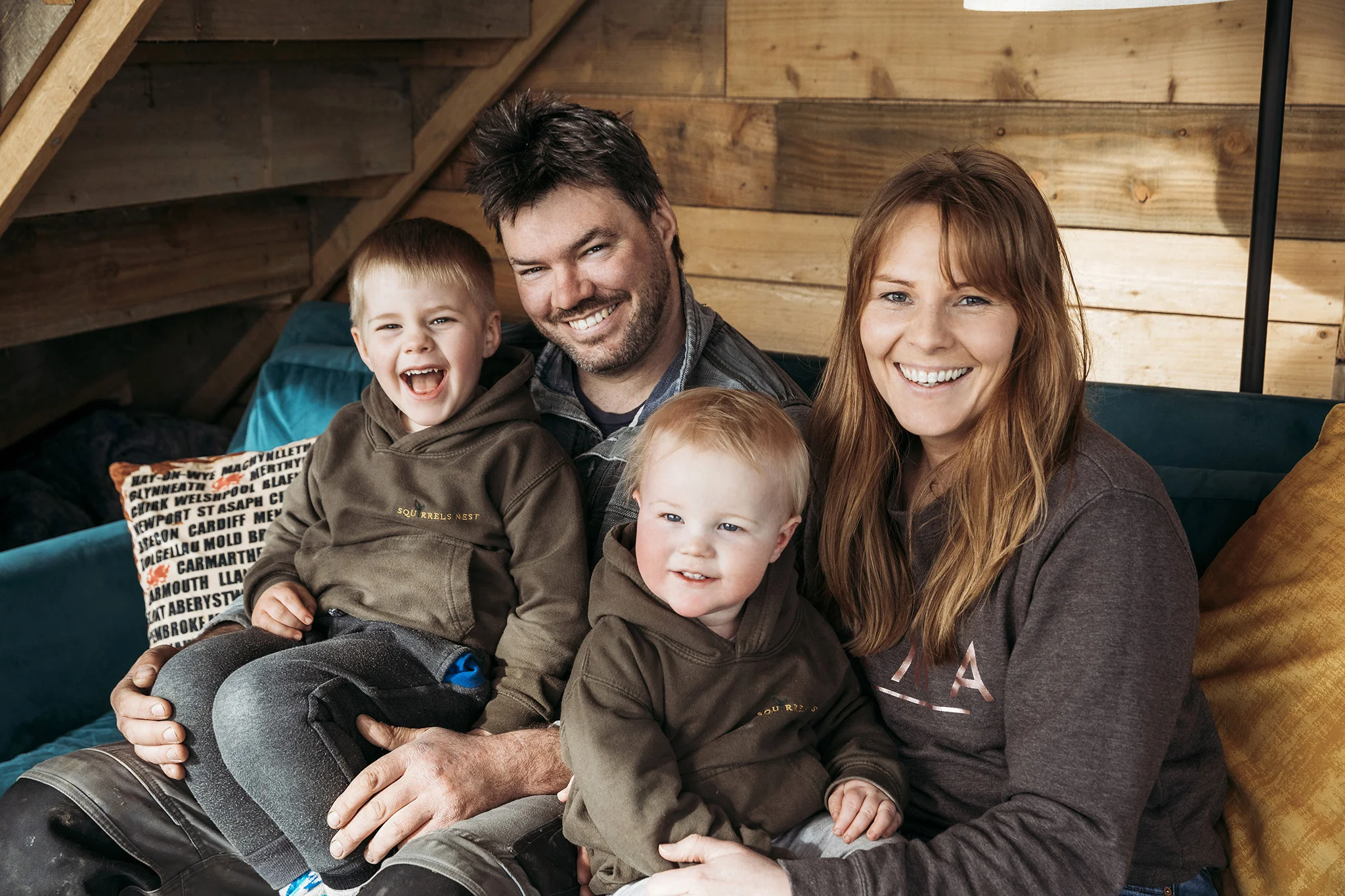 A family of four sits on a couch in a cozy wooden cabin. The parents are seated with their two young children, both smiling and wearing matching brown hoodies. The scene is warm and cheerful, like successfully logging in to the comfort of home, with the family cuddled closely together.