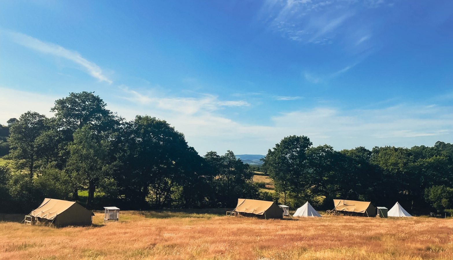 A serene campsite, known locally as Nantseren, features several canvas tents scattered across a golden grassy field. Tall trees and a lush forest border the field, with a clear blue sky and distant hills in the background. The area is bathed in sunlight, creating a peaceful outdoor setting.